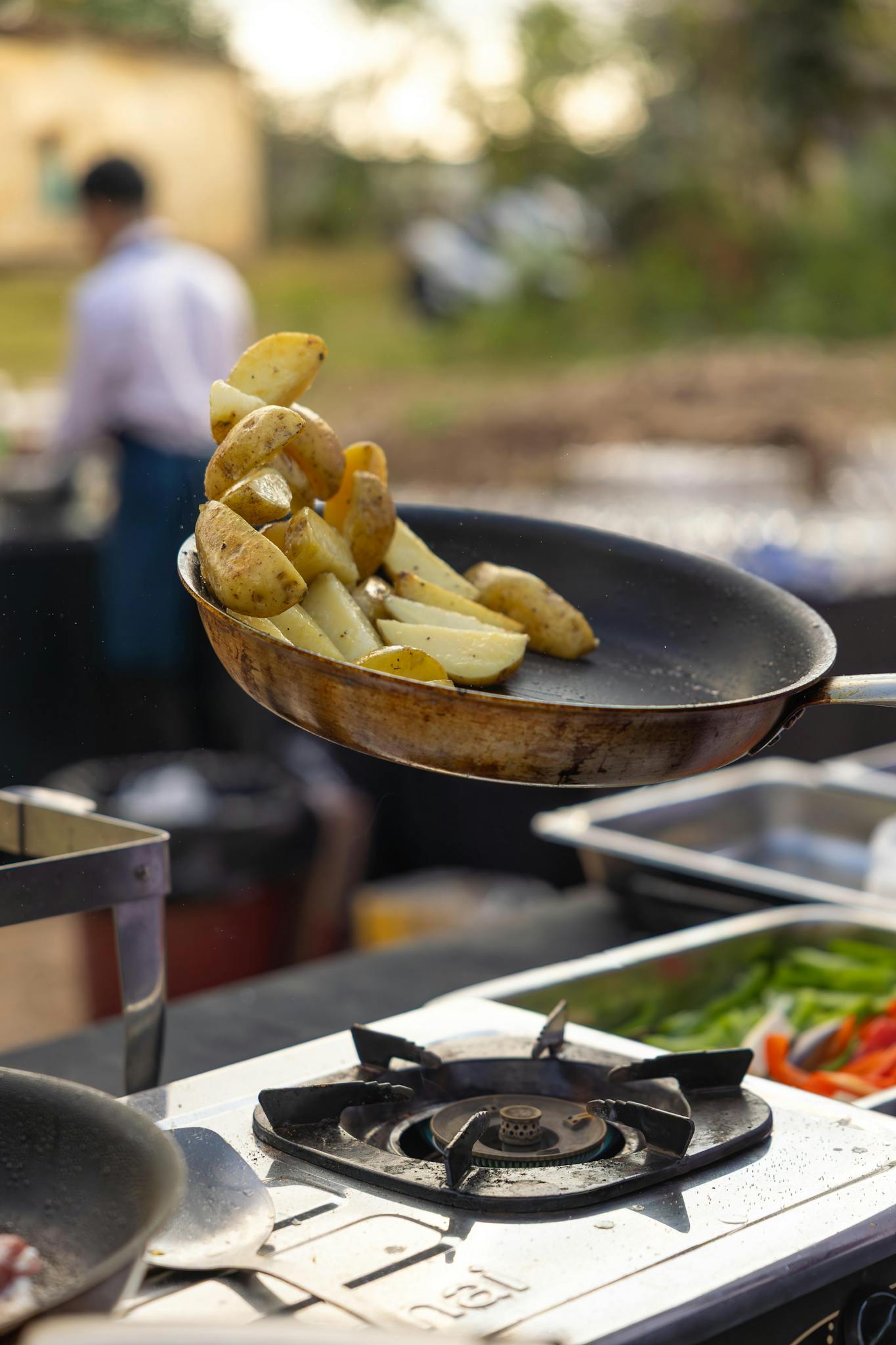 Chef tossing sautéed potatoes in pan at outdoor kitchen, showcasing culinary skills.