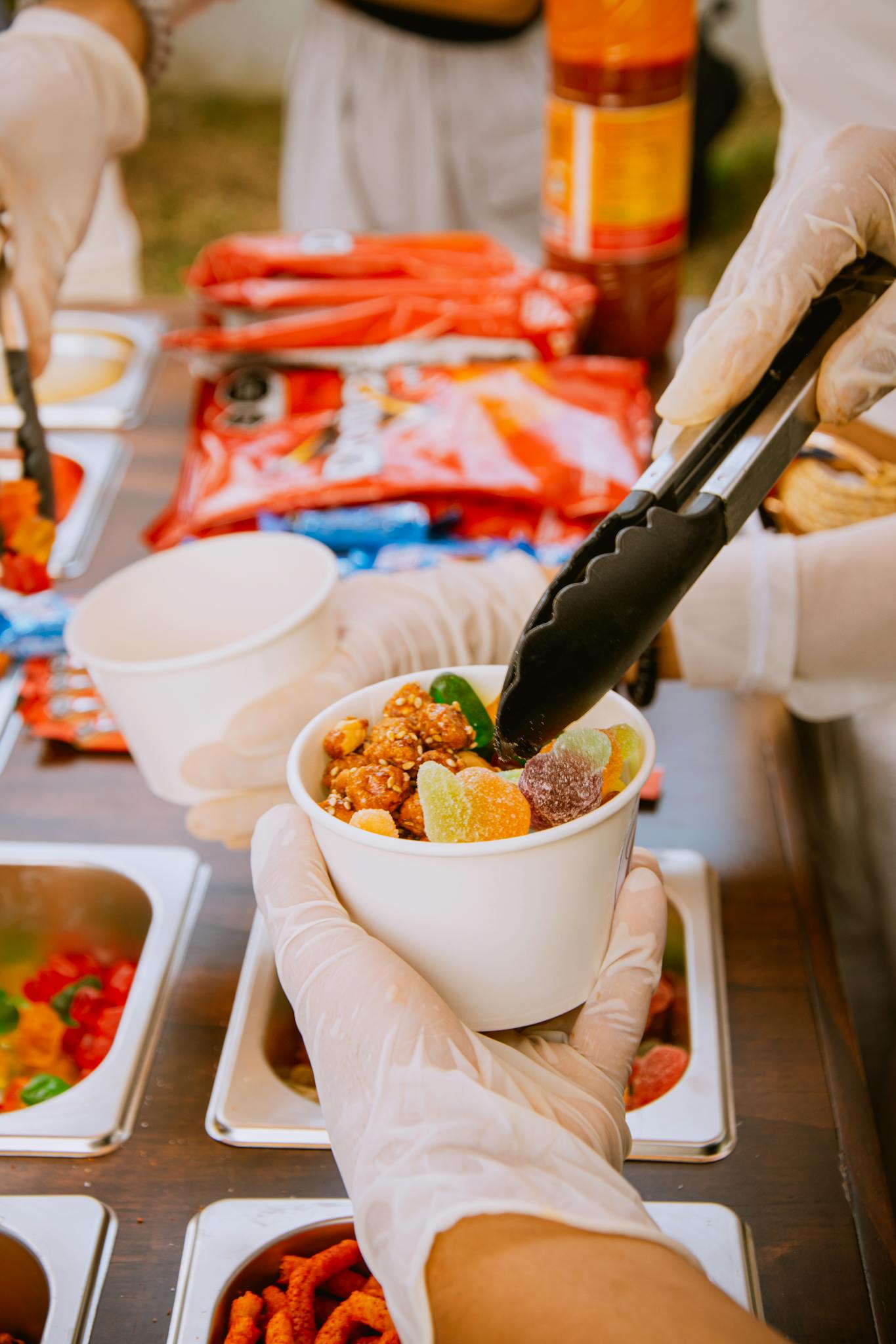 Gloved hands prepare a sweet snack with tongs, mixing colorful candies in a cup.